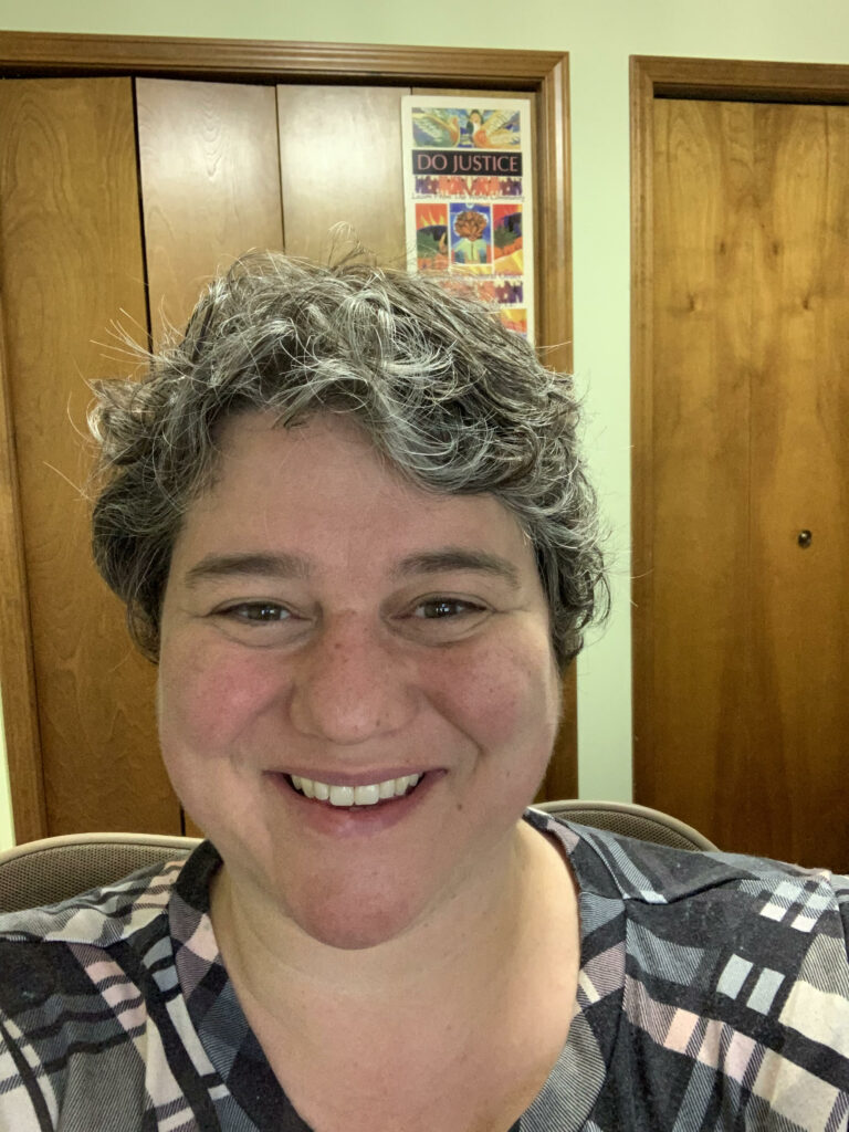 An image of a white woman with dark brown and gray curly short hair. The image is a selfie. She is sitting in her home office with a poster behind her that says "Do Justice'' she is wearing a black plaid vneck shirt.
