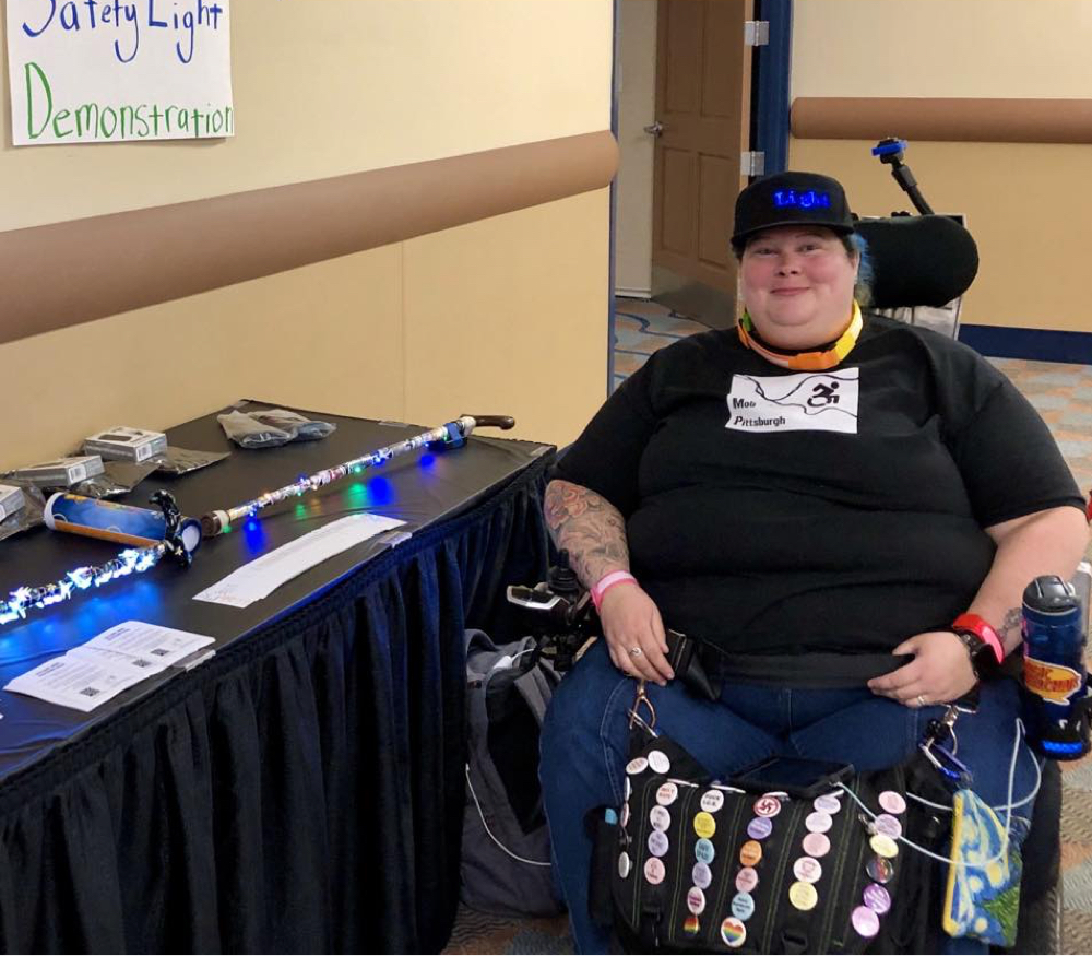 A person with pale ivory skin and rosy cheeks smiling while sitting in a power wheelchair . They are wearing a black hat and a black t-shirt. They are sitting in front of a table with papers on it.