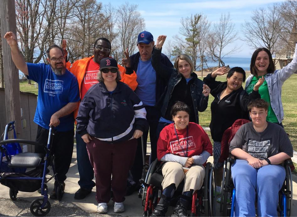A group of MDRC Program Participants posing for a picture. They are wearing multicolored t-shirts that read “Feisty &amp; Non-Compliant” Two people are in front of the other participant sitting in red and blue wheelchairs. Other participants stand behind them raising one hand and smiling.  