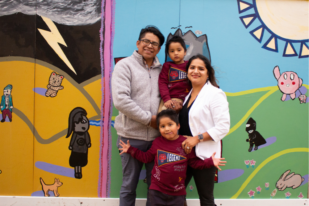 Picture of a family with caramel skin and dark brown hair; the tallest adult is wearing glasses with short hair and a light grey zip up sweater holding one child wearing a maroon shirt smiling. Second adult with long hair a black shirt and white blazer smiling with hand on shoulder of second child who is standing with a maroon shirt on. Family against a colorful mural.