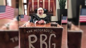 A smilinmg girl in her wheelchair in a judge's robe behind a large desk that reads "The Notorious RBG"