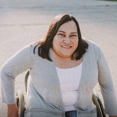 Woman sitting in wheelchair, with shoulder length brown hair, smiling