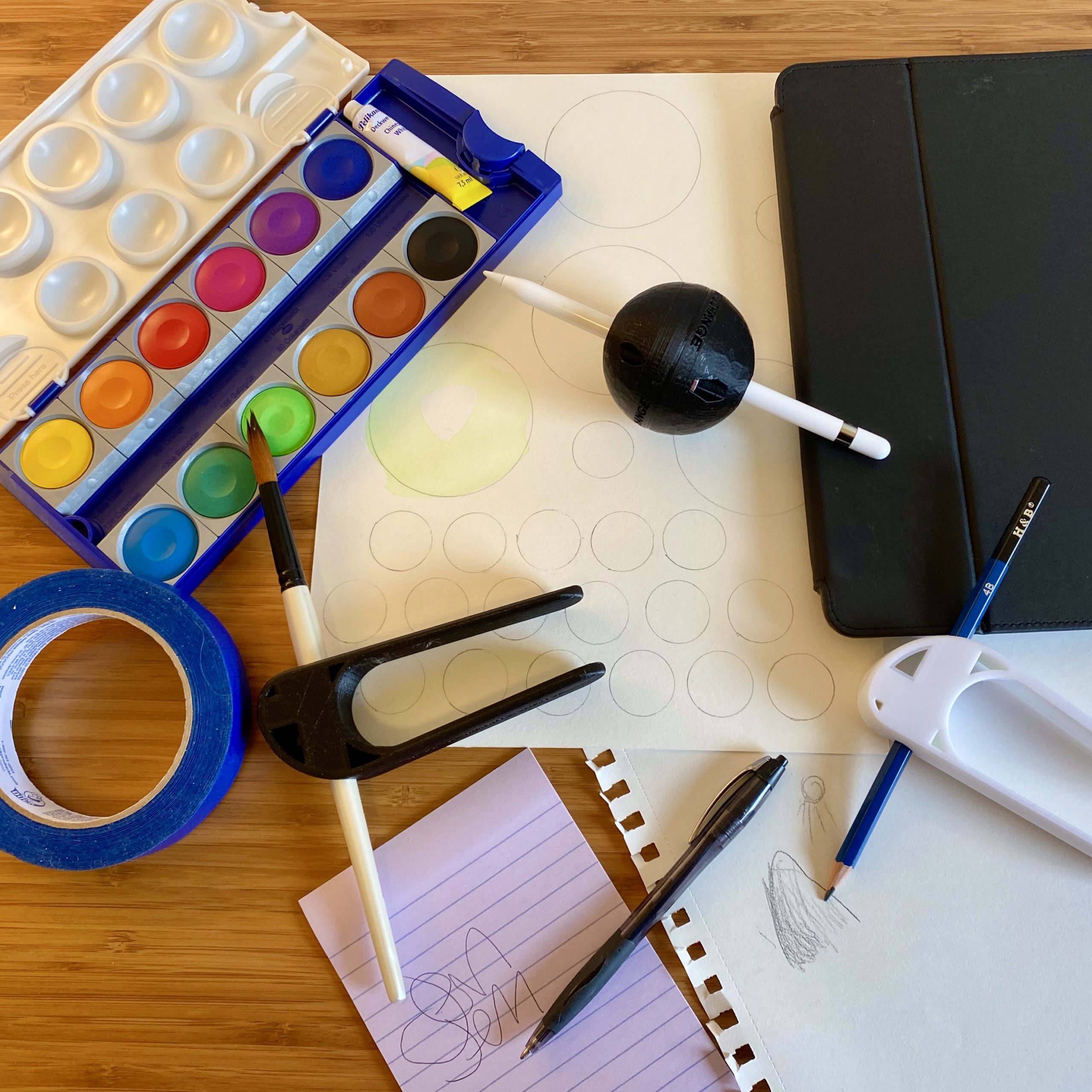 A watercolor paint palette, a Pen Ball device holding an Apple Pencil, a piece of watercolor paper, an iPad, a Palm Pen Holder device holding a blue drawing pencil pointing to a piece of sketch paper and small sketch of a hill and sunshine, a lined piece of paper with writing on it next to a pen, another Palm Pen Holder holding a paintbrush, and a roll of blue painters tape.  All of the items are on top of a wooden desk and the photo was taken from above.