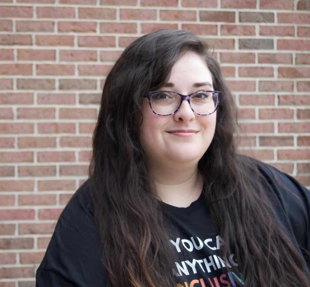 White woman of size, smiling with long wavy dark brown hair. Dark rimmed glasses and black t-shirt in background of photo is brick background