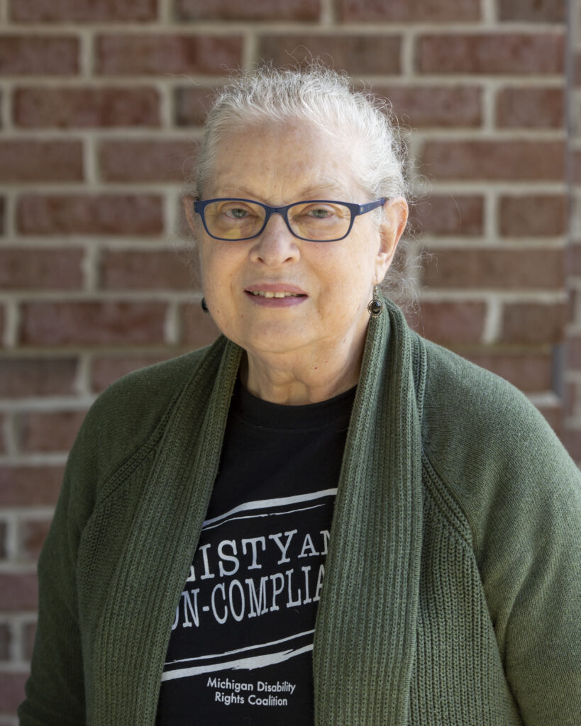 A white woman with gray and white hair pulled back in a ponytail is standing while smiling in front of a brick wall. She is wearing glasses with black frames and a black shirt with white text “Feisty and Non-Compliant”. Over her shirt she is wearing a dark green cardigan sweater.