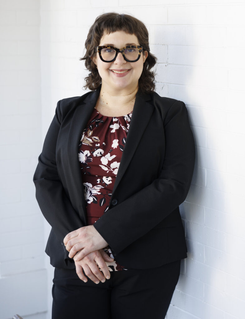 Woman wearing a suit jacket and fantastic glasses leaning against the wall, smiling.