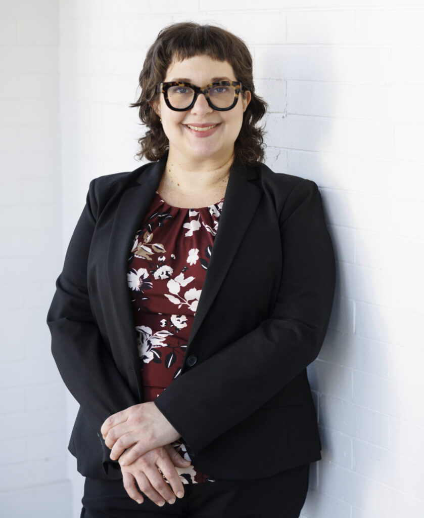 Woman wearing a suit jacket and fantastic glasses leaning against the wall, smiling.