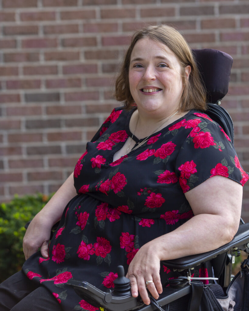A person with pale ivory skin and dark blonde hair smiling while sitting in a power wheelchair. They are wearing a black blouse with a pattern of red roses.