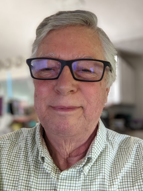 نورمان ديلايل ، رجل أبيض عجوز ، ذو شعر رمادي قصير ، يرتدي قميصًا أزرق متقلب بحمالات ؛ ابتسامة طفيفة. إنه جالس في مكتب الكمبيوتر الخاص به في المنزل ، مع جزء من مطبخ منزله في الخلفية.