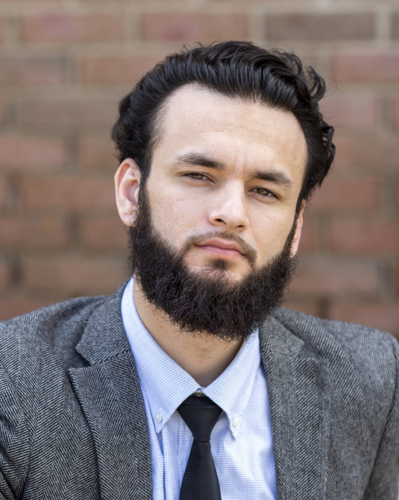 A person with tan skin, short black hair, and a black beard. They are wearing a gray suit jacket with a white and light blue collared button up shirt and a black tie. A brick wall is behind them.