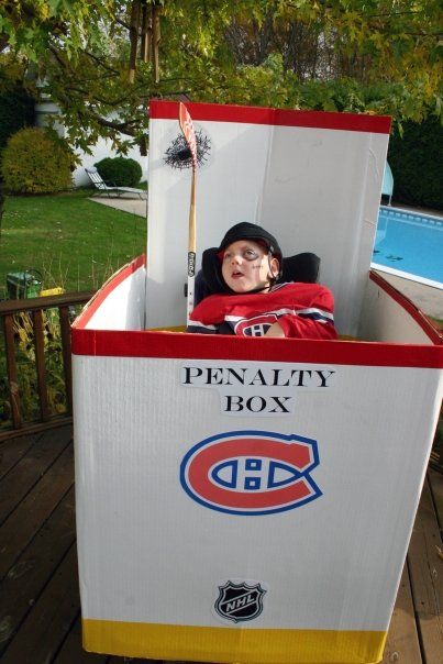 Young child in wheelchair wearing a hockey jersey and holding a hockey stick. His wheelchair is surrounded by a penalty box