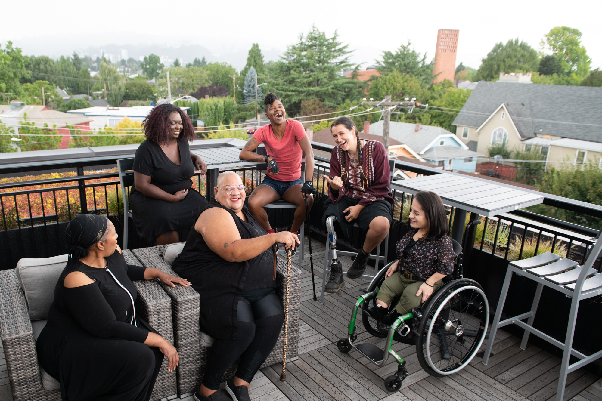 Overhead shot of six disabled people of color at a rooftop deck party. An Indigenous Two-Spirit person with a prosthetic leg smiles directly at the camera and gives a thumbs up while everyone else is engaged in conversation.  Photo from Disabled And Here.