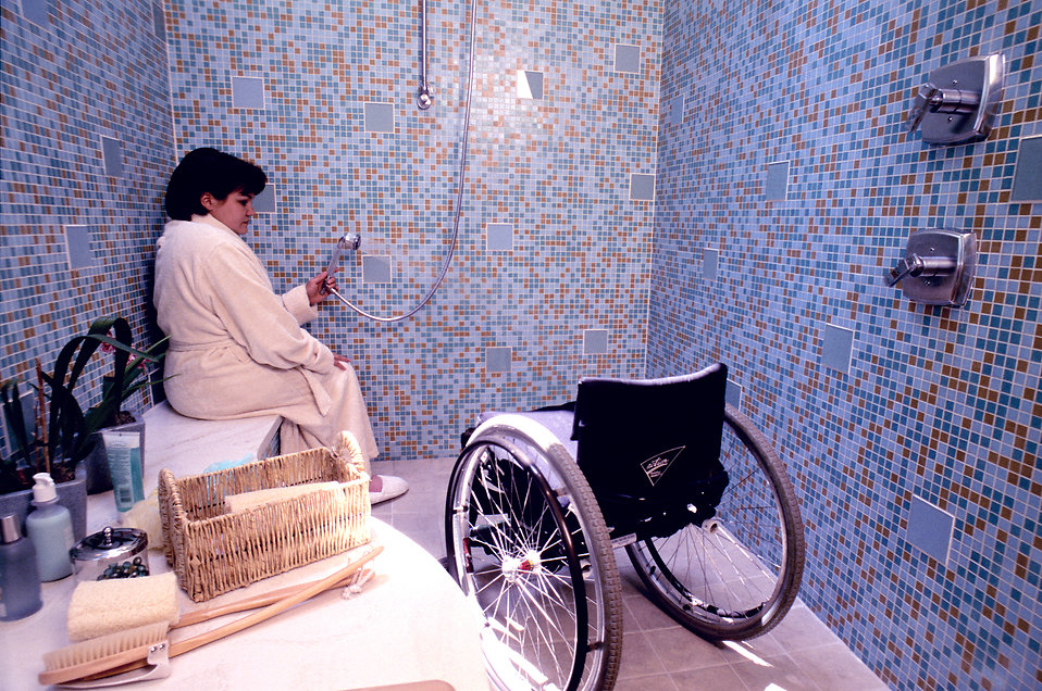 Woman seated in a shower, her wheelchair nearby.