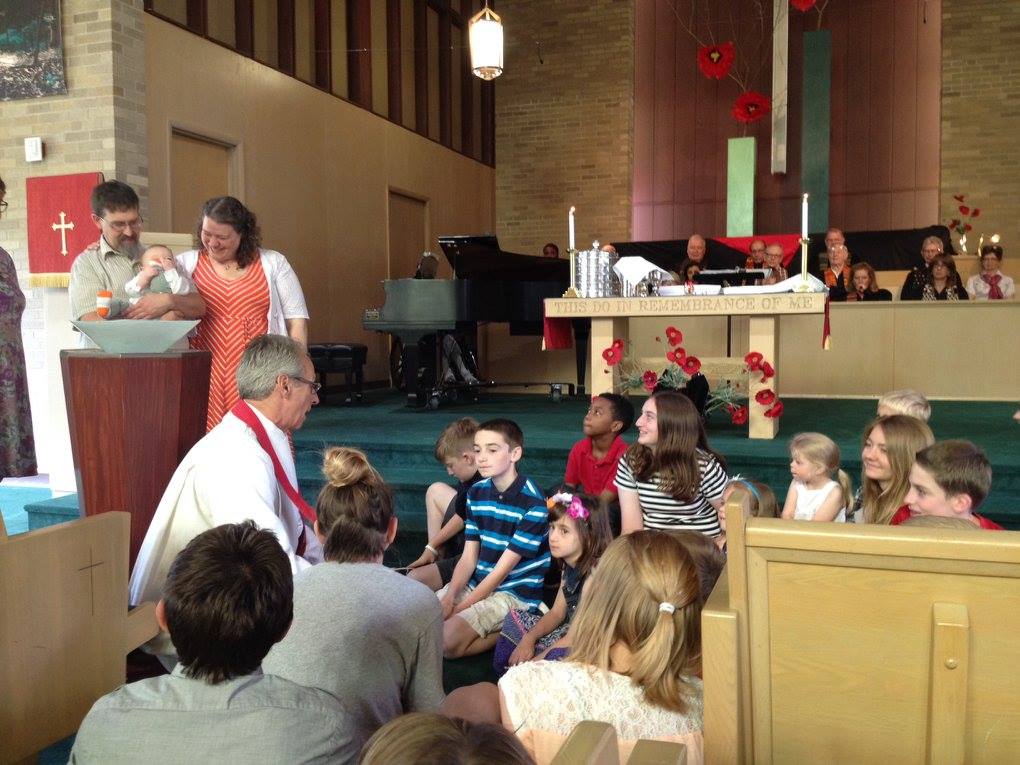Me next to my husband who is holding our son who is trying to eat his foot. We are next to the baptismal font. Our minister is crouched down in front of it talking to the congregation's children who are gathered around to hear about the meaning of baptism and to welcome Theo.