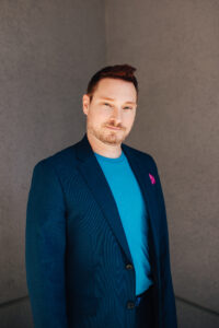  Headshot of Casey Stratton, a white man with short, reddish brown hair. Casey is wearing a dark blue blue blazer with a light blue T-shirt and magenta pocket square and is standing in front of a dark grey wall