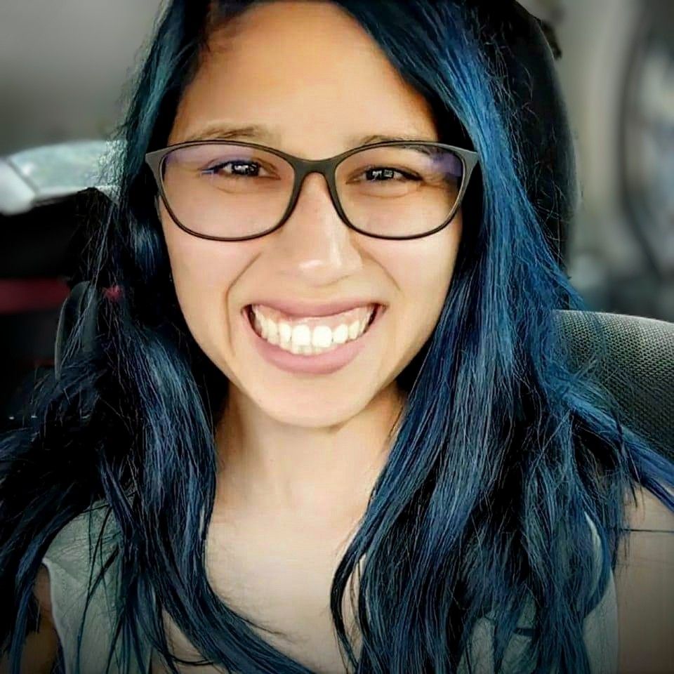 [Image description: a latina woman with long dark blue hair. Priscilla is looking directly at the camera, smiling. She is wearing black framed glasses. End of image description.]