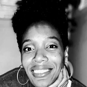 Black and white headshot of Feliece Turner, a Black woman with short black hair in an afro style. Feliece is sitting on steps with a plant behind her. Feliece is facing the camera with one hand on her chin and is smiling. She is wearing a t-shirt, cardigan and silver hoop earrings