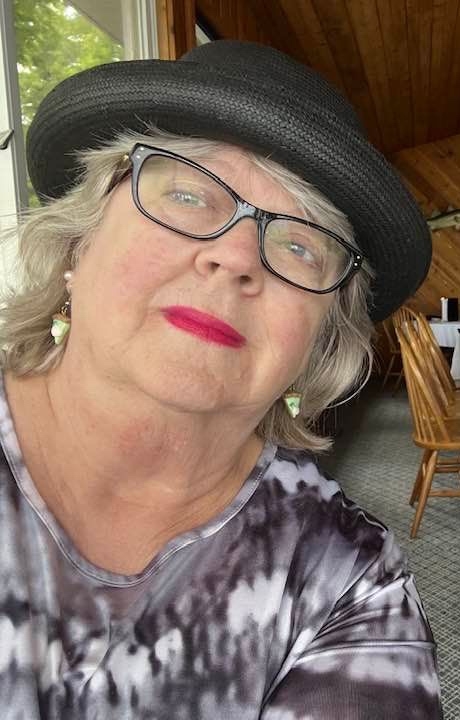 An older white woman with gray hair who is wearing black glasses, a black and white tie-dye shirt, a black hat and pink lipstick. She is sitting in a waterfront restaurant, gazing thoughtfully at a lake in northern Michigan.