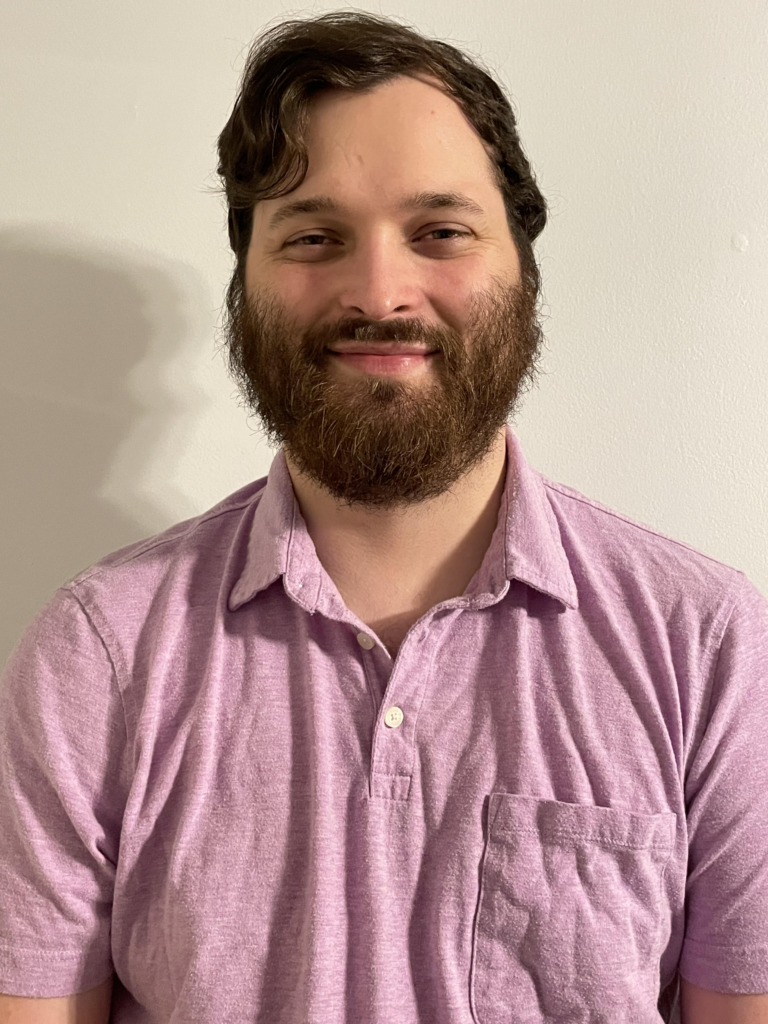 [Image description: Headshot of Brandon Taylor, a white man with short brown hair and a beard. Brandon is standing inside, with a blank white wall behind him. He is looking directly at the camera, smiling, and is wearing a light purple polo shirt. End image description.]